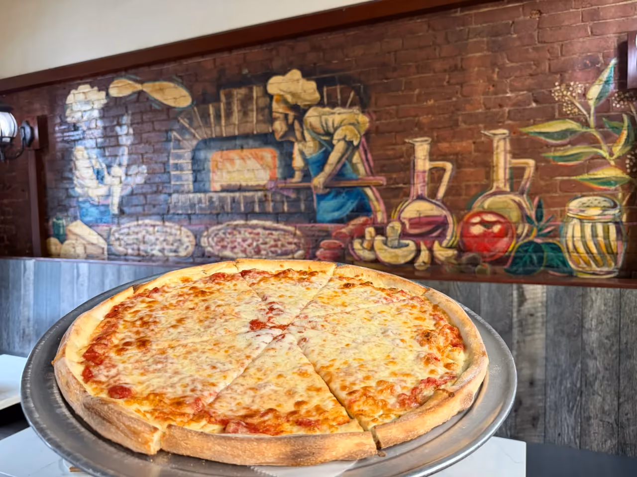A whole cheese pizza on a tray in front of Antonio's pizzaiolo mural