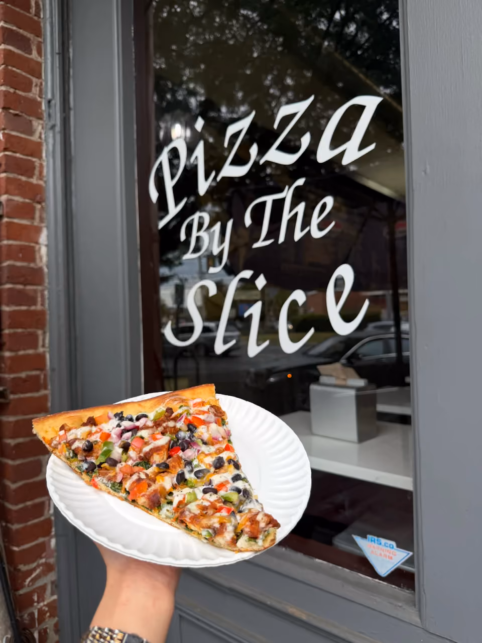 A loaded gourmet slice held at Antonio's Pizza By The Slice window